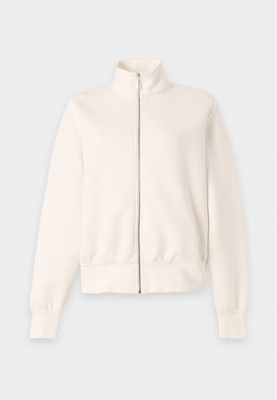 Veste zippée à manches longues blanc cassé avec col montant et poignets et bas côtelés sur fond gris clair.