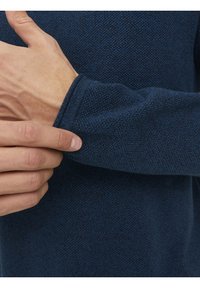 Personne ajustant le poignet d'un pull à manches longues bleu foncé texturé, montrant le détail du tissu tricoté et la position de la main.