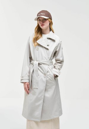 Jonge vrouw die een lichtgrijze trenchcoat met ceintuur en een bruine baseballpet draagt, staand met één hand in de zak tegen een effen achtergrond.