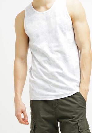 Hombre con camiseta sin mangas blanca con estampado y pantalones cortos cargo verdes oscuros con una mano en el bolsillo.