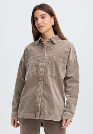 Kvinde med langt brunt hår iført beige oversized skjorte og matchende bukser, stående mod en ensfarvet lys baggrund.