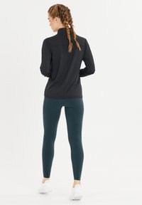 Musta urheilu takki, jossa on korkea kaulus, yhdistettynä teal-värisiin legginsseihin. Legginsseissä on sivutaskut ja ne on yhdistetty valkoisiin urheilukenkiin.