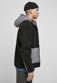 Jachetă din fleece neagră cu glugă gri, având fermoar frontal, țesătură texturată și accente cu petice pe laterale. Croi standard cu mâneci lungi.