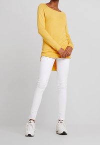 Top amarillo de manga larga con escote barco, combinado con jeans ajustados blancos y zapatillas blancas. El top cuenta con un dobladillo asimétrico.