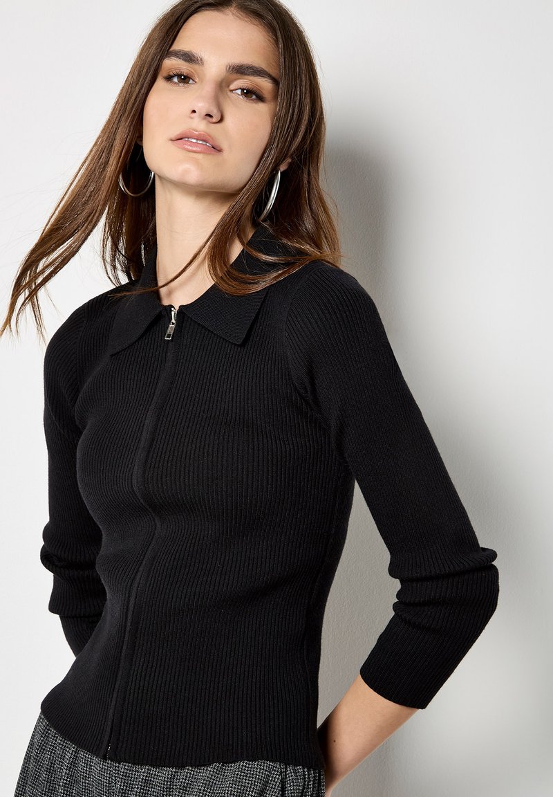 Top in maglia a coste nera con zip frontale e ampio colletto. Maniche lunghe con polsini aderenti. Tessuto leggero con una texture liscia.