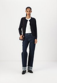 Black cardigan, white ribbed top, dark blue wide-leg jeans with logo cuffs, and black heels. Model carries a small brown handbag.