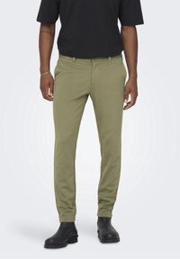 Pantalon sur mesure vert olive en tissu extensible, avec poches avant, passants de ceinture et coupe slim, associé à des bottines noires.