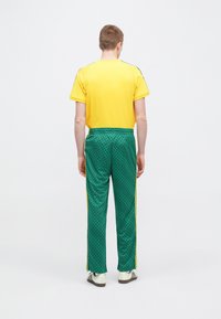 Gelbes kurzärmliges Hemd mit grünen, gemusterten Trainingshosen mit Seitenstreifen und lockerem Sitz. Das Outfit wird mit gestreiften Turnschuhen kombiniert.