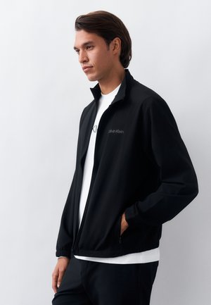 Άνδρας που φοράει μαύρο ζακέτα με φερμουάρ Calvin Klein πάνω από λευκό μπλουζάκι, στέκεται με το ένα χέρι στην τσέπη, κοιτώντας προς τα πλάγια.