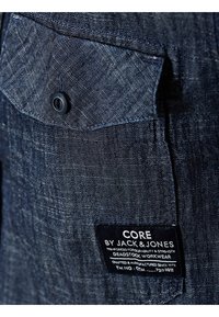 Dżinsowa koszula z niebieskiej teksturowanej tkaniny, z ostrym kołnierzykiem i patką z kieszenią zapinaną na czarny guzik. Metka z napisem „CORE by JACK & JONES”.
