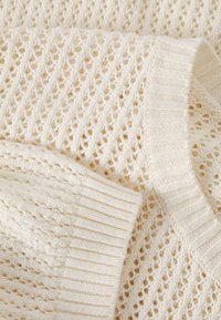 Il tessuto lavorato a maglia color crema presenta un motivo a tela aperta e bordi a coste, mettendo in risalto un pattern testurizzato e un design leggero.