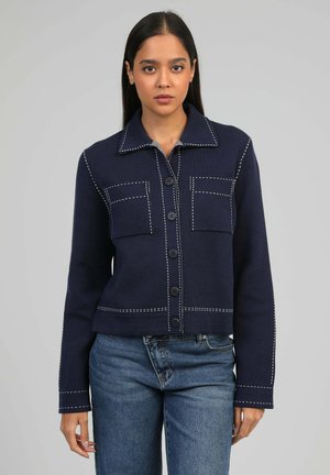 Femme portant une veste bleu marine boutonnée avec des surpiqûres blanches et des poches avant, associée à un jean bleu, faisant face à un fond uni.