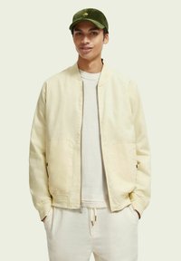 Helle beige Bomberjacke mit zwei vorderen Taschen, getragen über einem hellen Pullover. Eine dunkelgrüne Cordmütze ziert ein gelbes gesticktes Design.