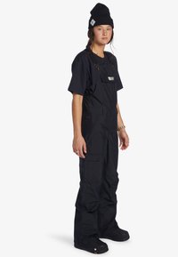 Mladá osoba v černé čepici, černém tričku s krátkým rukávem, černých lacláčích s kapsami a černých botách, stojící s mírným pohledem vlevo.