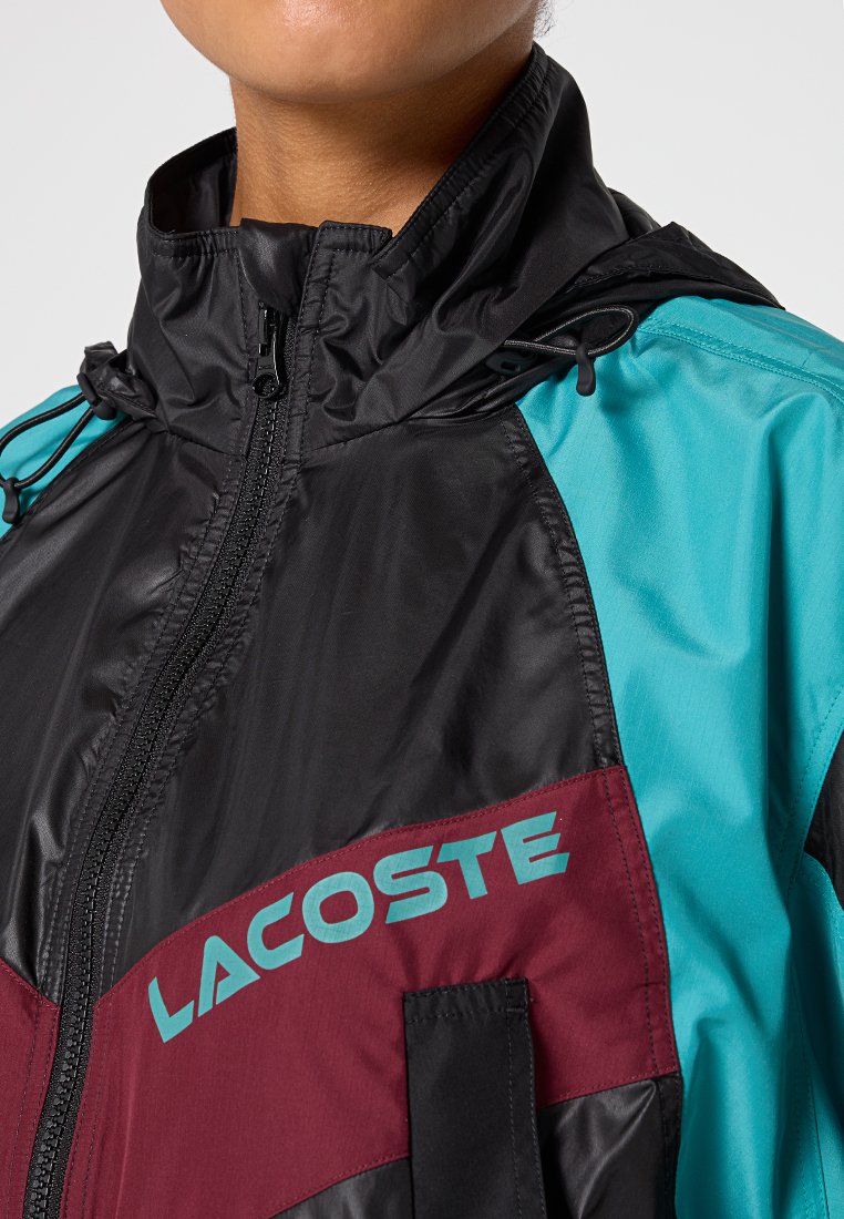 Zbliżenie na osobę w kurtce przeciwwiatrowej na zamek, w czarne, turkusowe i bordowe panele, z napisem "LACOSTE" na klatce piersiowej.