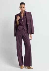 Paarse op maat gemaakte pak met een cropped blazer, gouden knopen en een wijduitlopende broek. Vergezeld van huidskleurige hakken en opvallende gouden oorbellen.