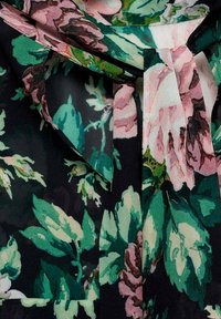 Blouse florale avec un fond noir, ornée de grandes fleurs roses et vertes, en tissu doux et avec un détail noué au niveau du col.