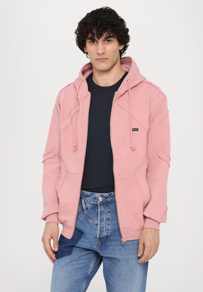 Rosa Zip-Pullover mit strukturiertem Design, ausgestattet mit einer Kapuze mit Kordelzug und Kängurutaschen, getragen über einem marineblauen Shirt und hellblauen Jeans.