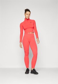 Lysende korallfarget treningstøy bestående av en tettsittende langermet topp med høy krage og matchende leggings. Begge har logoaksenter.