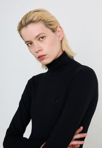 Sort rullekrave sweater med ribbet tekstur, slim fit, med et lille logo på brystet. Modellen har lyst blont hår.