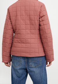 Prošívaná růžová bunda s dlouhými rukávy, zakulacenými rameny a úzkým pasem. Kombinována s volnými džínami, které ukazují uvolněný střih.