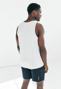 Weißes ärmelloses Sportshirt mit strukturierter Oberfläche, kombiniert mit schwarzen Shorts, die einen seitlichen Schlitz und reflektierende Akzente aufweisen.