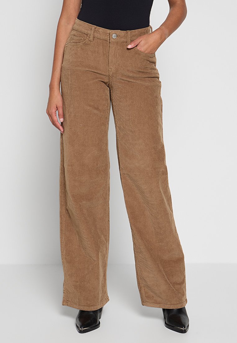 Braune Corduroy-Weitbundhosen mit Knopfverschluss, Fronttaschen und einer weichen Textur. Kombiniert mit einem enganliegenden Oberteil und schwarzen Stiefeln.