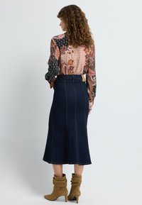 Niebieska dżinsowa spódnica midi z widocznym szwem, w połączeniu z kolorową bluzką w wzór paisley i beżowymi, luźnymi botkami za kostkę.