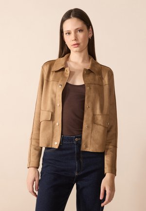 Veste cropped en suédine marron avec un col, deux poches avant, des boutons-pression et des coutures contrastantes, assortie à un jean foncé et un haut marron.