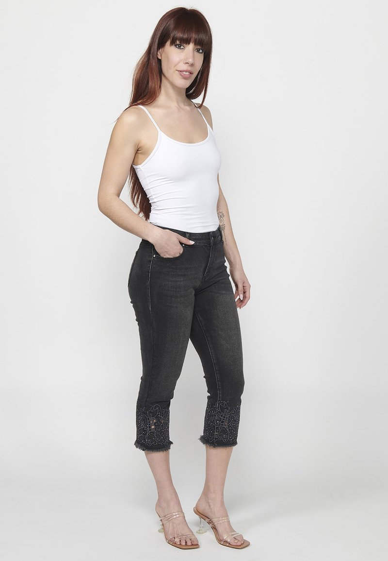 Crne traperice visokog struka s izvezenim rubovima i strganim rubovima, u kombinaciji s bijelom majicom bez rukava i prozirnim sandalama s potpeticom.