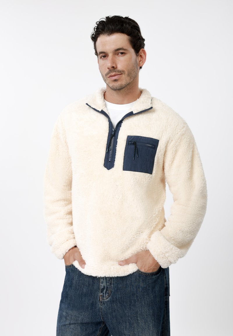 Φλις σε κρεμ χρώμα με σχέδιο με μισό φερμουάρ, μπλε τσέπη στο στήθος και υφή με ανάγλυφο ύφανμα. Διαθέτει ελαστικά μανίκια και ρεβέρ.