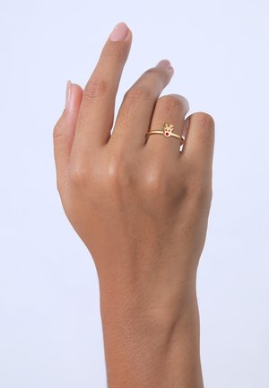 Goldene Metallring mit einem kleinen Rentierdesign, das mit einem roten Schmuckstein akzentuiert ist, getragen an einer Hand mit heller Haut und neutraler Nagelfarbe.