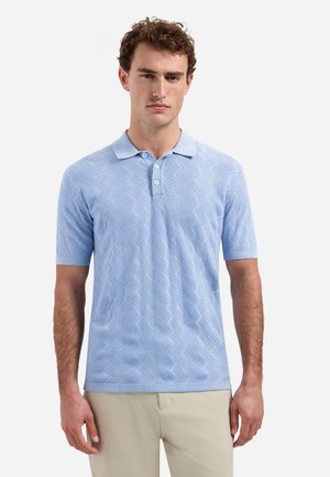 Man draagt een lichtblauw, gestructureerd gebreid poloshirt en beige broek, staand tegen een effen witte achtergrond.