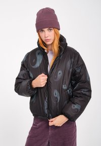 Giacca a vento nera con un motivo yin-yang in tonalità di teal, con colletto alto e zipper frontale. Indossata con un berretto di maglia viola.
