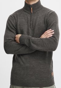 Szary sweter dzianinowy z wysokim kołnierzem, ćwierć zamkiem, ściągaczami przy mankietach i dole oraz małą skórzaną naszywką z logo z boku.
