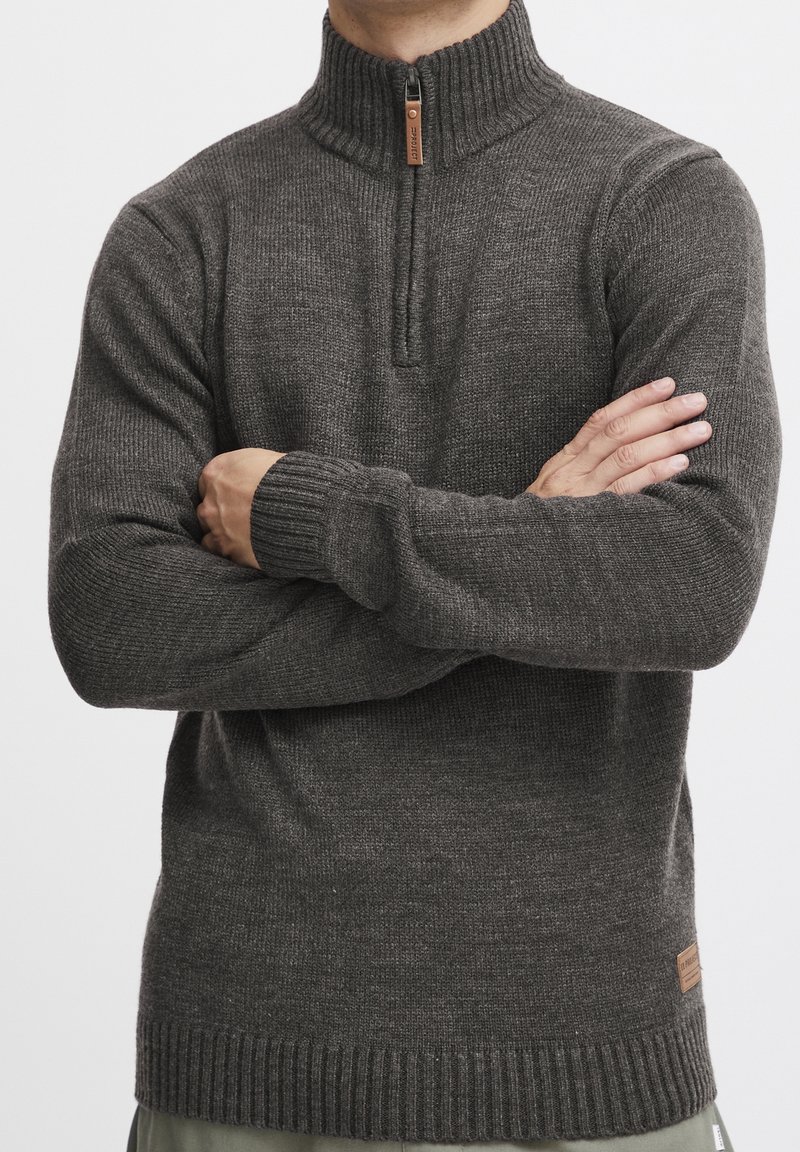 Szary sweter dzianinowy z wysokim kołnierzem, ćwierć zamkiem, ściągaczami przy mankietach i dole oraz małą skórzaną naszywką z logo z boku.