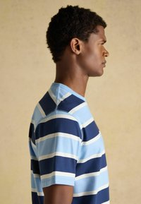 Jeune homme de profil portant une chemise à manches courtes à larges rayures horizontales bleu clair, bleu marine et blanc sur un fond uni.