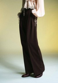 Pantalons marron à jambes larges avec une texture lisse, associés à un haut clair doté de volants et de liens aux poignets, portés avec des bottes foncées.