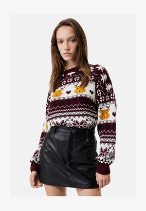 Jeune femme portant un pull à motifs de rennes bordeaux, blanc et jaune et une mini-jupe en cuir noire, debout avec une expression neutre.