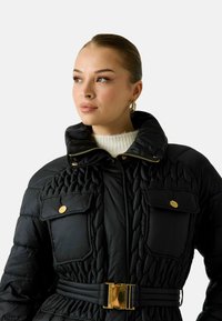 Giacca imbottita nera con collo alto, bottoni dorati, texture trapuntata, vita con cintura e due tasche frontali.