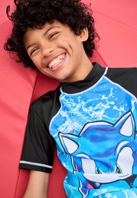 Kortærmet badetrøje med en blå Sonic the Hedgehog-grafik mod et vandsprøjtmønster på sort baggrund; båret af et smilende barn.