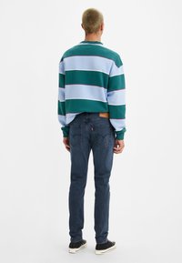 Grüner und hellblauer gestreifter Sweatshirt mit rundem Ausschnitt, kombiniert mit dunkelblauen Jeans und schwarzen Sneakers. Dezentes Logo auf den Jeans.