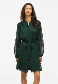 Rochie verde cu mâneci lungi din material transparent �și talie accentuată cu cureaua. Are un tiv cu volane și o textură de material neted.