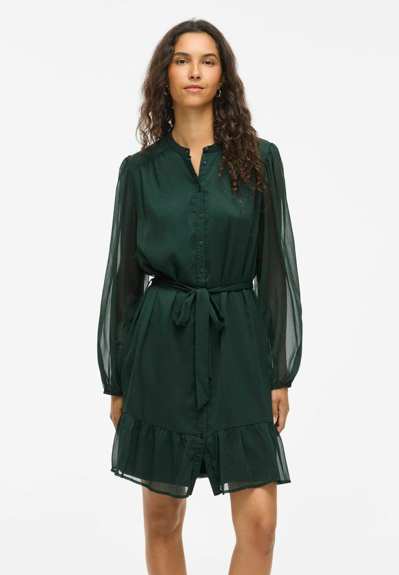 Rochie verde cu mâneci lungi din material transparent și talie accentuată cu cureaua. Are un tiv cu volane și o textură de material neted.