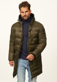 Olivgrüne Steppjacke mit Kapuze, Reißverschluss und horizontalen Steppnähten; getragen über einem marineblauen Rollkragenpullover und hellen Jeans.