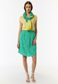 Gelber gestrickter Oberteil kombiniert mit einem grünen, gemusterten Rock. Das Outfit umfasst einen grünen und gelben gemusterten Schal und weiße Sandalen mit Riemchen.