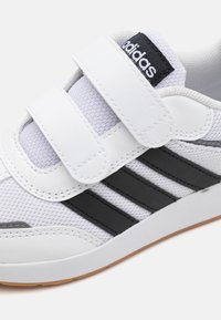 Hvit atletisk sko med svarte striper, borrelåsremmer, mesh-stoff og gummisåle. "adidas"-logo på tungen; glatte teksturdetaljer.