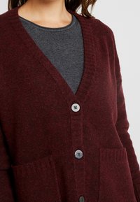 Bordowy wełniany kardigan z dekoltem w serek, z zapięciem na guziki, dwoma przednimi kieszeniami i miękkim, teksturowanym wykończeniem.