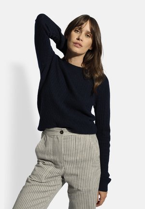 Marineblauer Strickpullover mit gerippter Textur, Rundhalsausschnitt und langen Ärmeln, kombiniert mit gestreiften, hochgeschnittenen Hosen mit Knopfdetails.