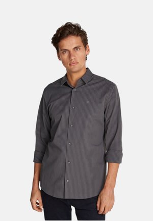Uomo che indossa una camicia slim fit grigio scuro a maniche lunghe con sottili righe verticali e maniche risvoltate su uno sfondo semplice.